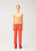 A woman with light skin and red hair in a ponytail models the MONTHLY COLORS : SEPTEMBER collection by PLEATS PLEASE ISSEY MIYAKE, wearing a short-sleeved, ribbed orange top paired with red straight pants and lace-up boots. She stands against a plain light background, her ensemble’s color palette creating a striking yet harmonious effect.