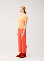 A woman with light skin and long red hair, dressed in the PLEATS PLEASE ISSEY MIYAKE MONTHLY COLORS : SEPTEMBER collection—including a yellow short-sleeve top and orange straight pants—stands in profile against a plain light background. The gentle tones of her outfit from this collection create a harmonious color composition.