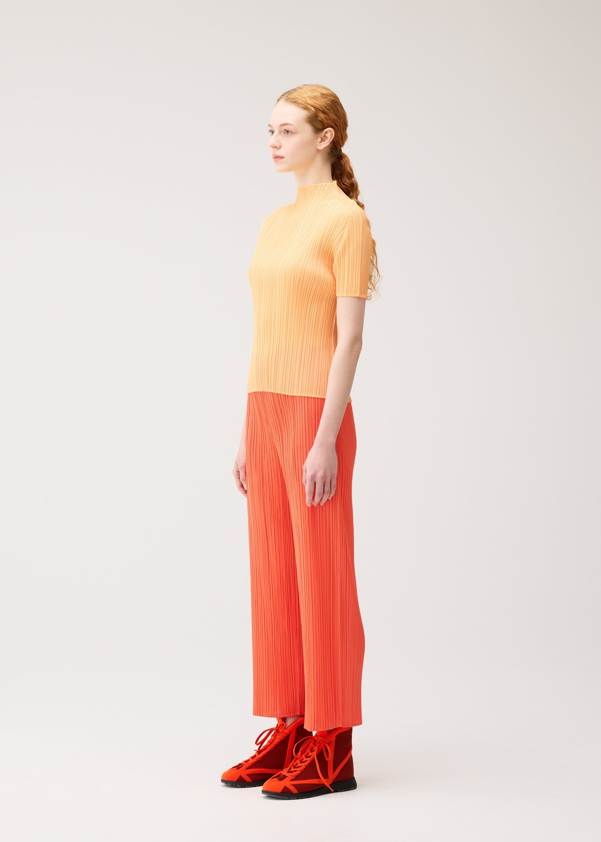 Zoomed view A woman with light skin and long red hair, dressed in the PLEATS PLEASE ISSEY MIYAKE MONTHLY COLORS : SEPTEMBER collection—including a yellow short-sleeve top and orange straight pants—stands in profile against a plain light background. The gentle tones of her outfit from this collection create a harmonious color composition.