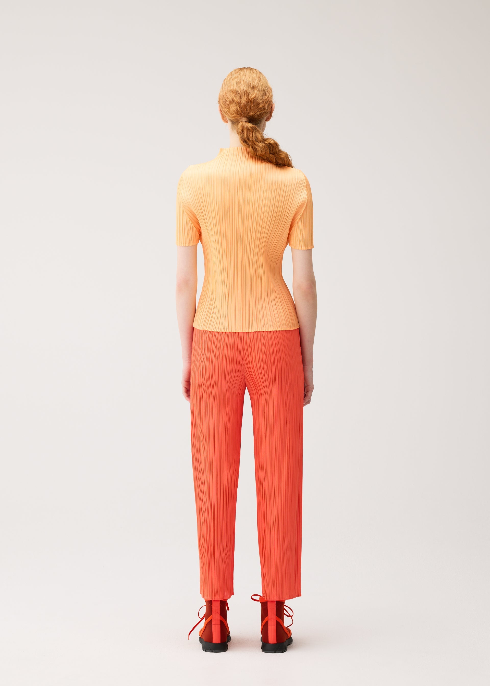 Zoomed view Wearing pieces from the PLEATS PLEASE ISSEY MIYAKE collection, MONTHLY COLORS : SEPTEMBER, a person with long red hair in a low ponytail faces away against a plain light background. Their ensemble features a short-sleeved, ribbed light orange top paired with bright orange pleated pants and black shoes. The harmonious image is enhanced by gentle tones and balanced color composition.