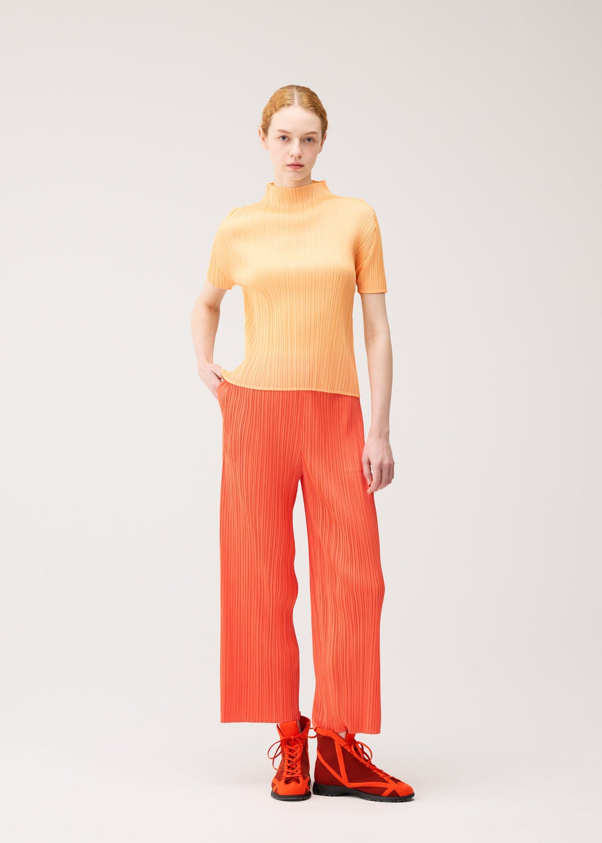 A woman with light skin and red hair tied back stands against a plain background, wearing the PLEATS PLEASE ISSEY MIYAKE MONTHLY COLORS : SEPTEMBER ensemble—an orange short-sleeve top and matching straight pants paired with orange shoes featuring black soles. With one hand in her pocket, she creates a striking color composition that draws the eye. view 10 of 10
