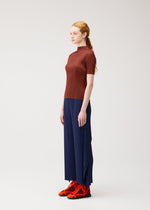 A woman with red hair in a low ponytail models pieces from PLEATS PLEASE ISSEY MIYAKE’s MONTHLY COLORS: SEPTEMBER collection, featuring a short-sleeved brown top and wide-leg navy pleated flare pants. Paired with vibrant orange shoes and set against a plain light background, the look artfully balances gentle tones with bold accents for a harmonious yet striking ensemble.