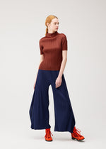 A person with red hair wears the MONTHLY COLORS : SEPTEMBER ribbed, short-sleeve brown top and wide-leg navy flare pants by PLEATS PLEASE ISSEY MIYAKE, paired with bright orange lace-up shoes, standing against a plain light background. The ensemble from PLEATS PLEASE ISSEY MIYAKE highlights an appealing color composition that draws the eye in gentle tones.