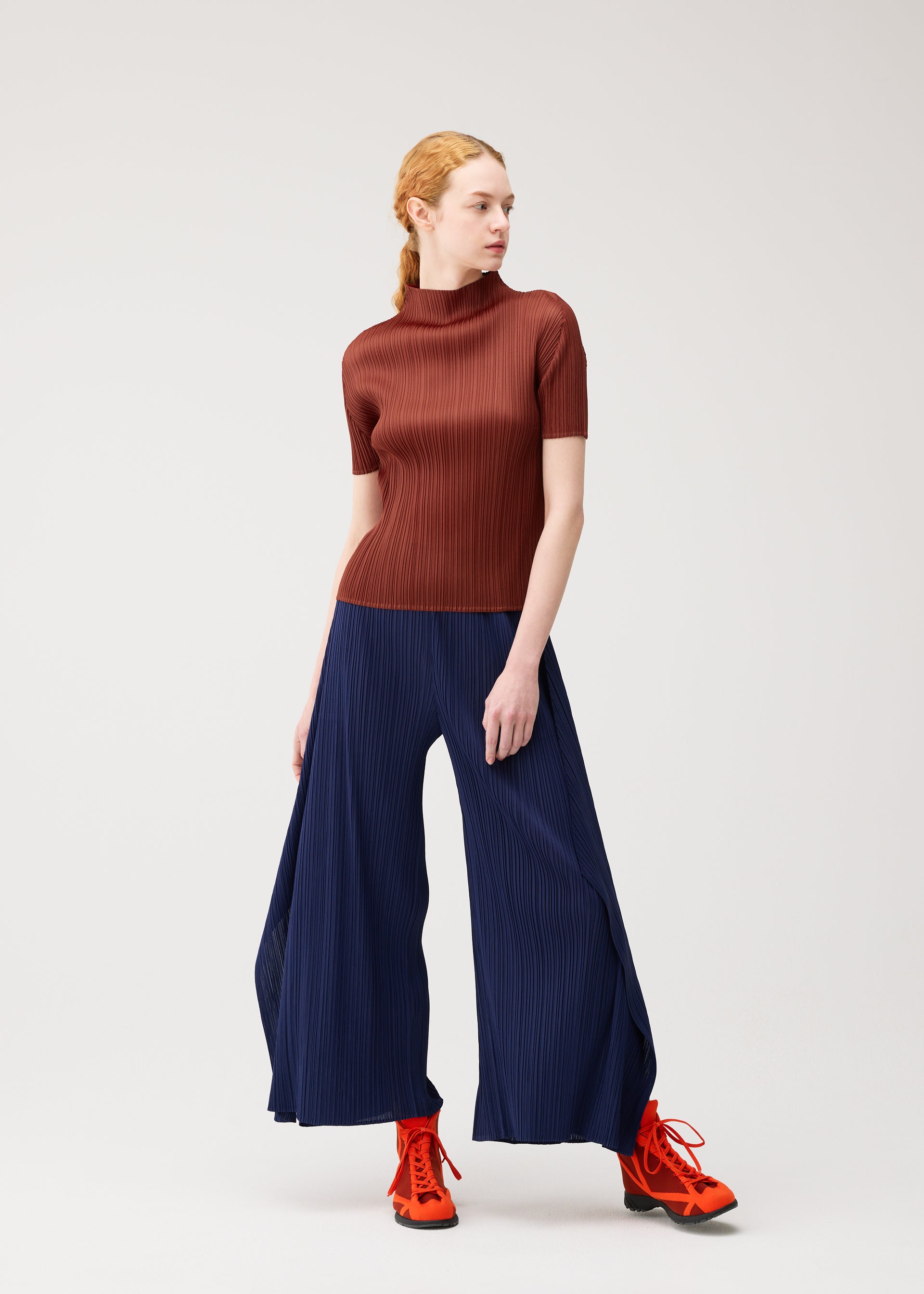 Zoomed view A person with red hair wears the MONTHLY COLORS : SEPTEMBER ribbed, short-sleeve brown top and wide-leg navy flare pants by PLEATS PLEASE ISSEY MIYAKE, paired with bright orange lace-up shoes, standing against a plain light background. The ensemble from PLEATS PLEASE ISSEY MIYAKE highlights an appealing color composition that draws the eye in gentle tones.