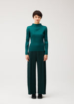 A person stands facing forward in the MONTHLY COLORS : OCTOBER teal pleated top and matching wide pants by PLEATS PLEASE ISSEY MIYAKE, paired with black shoes against a plain light background.