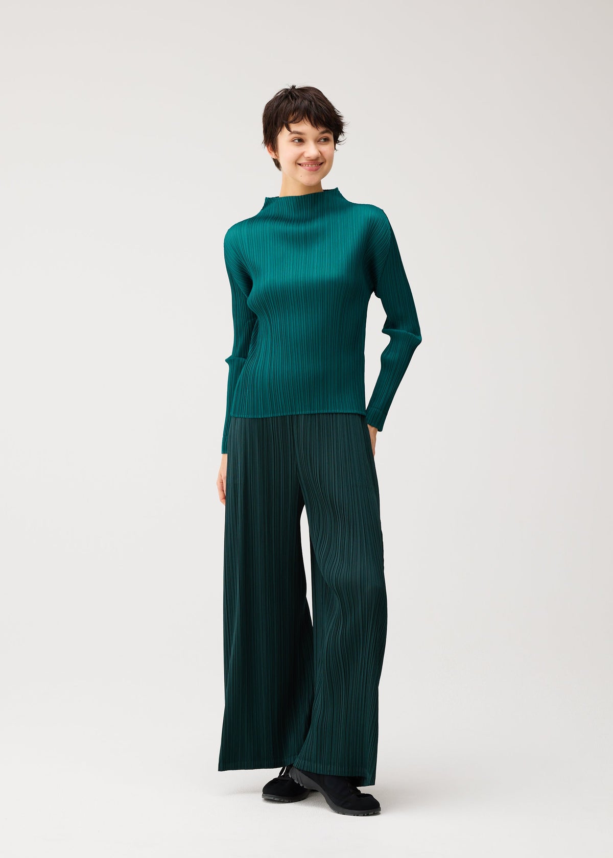 A person stands against a plain background, smiling and looking to the side, wearing the teal MONTHLY COLORS : OCTOBER set by PLEATS PLEASE ISSEY MIYAKE with black shoes. view 10 of 10