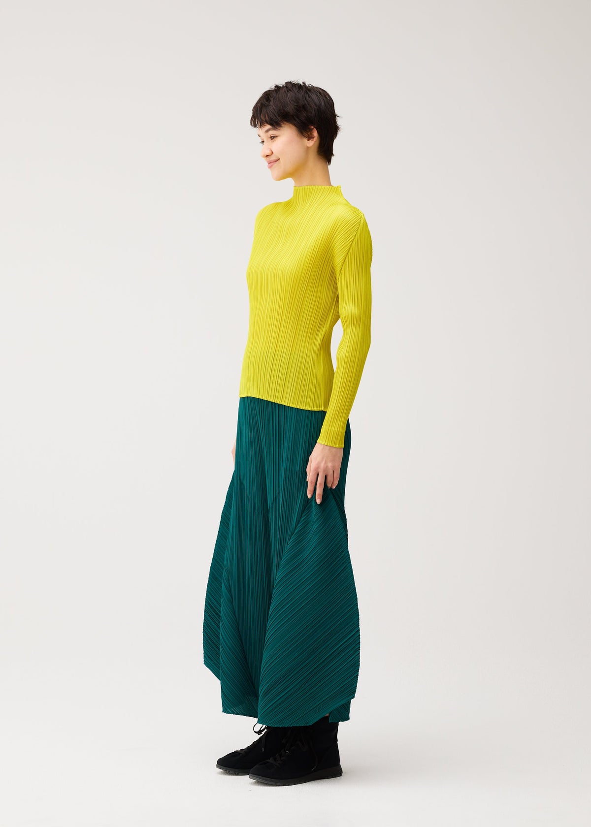A woman with short dark hair smiles sideways, wearing the MONTHLY COLORS : OCTOBER by PLEATS PLEASE ISSEY MIYAKE—a bright yellow ribbed turtleneck sweater and flared pants against a plain white background. view 9 of 11