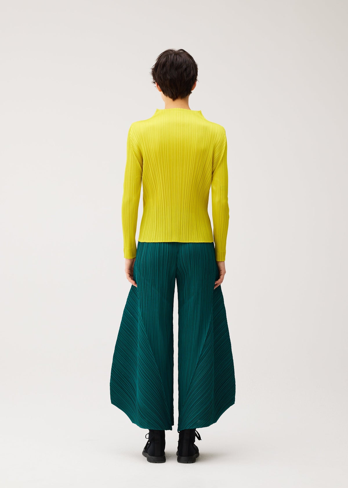 Zoomed view A person stands with their back to the camera, wearing MONTHLY COLORS : OCTOBER wide-legged teal color block pants by PLEATS PLEASE ISSEY MIYAKE and a yellow long-sleeve top against a plain light background.