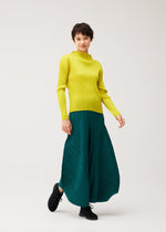 A person in MONTHLY COLORS : OCTOBER by PLEATS PLEASE ISSEY MIYAKE stands smiling, one foot forward in a yellow long-sleeve top and teal pleated skirt against a plain light background.