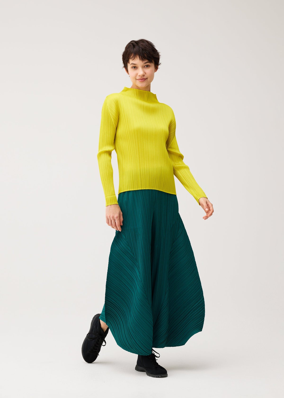 Zoomed view A person in MONTHLY COLORS : OCTOBER by PLEATS PLEASE ISSEY MIYAKE stands smiling, one foot forward in a yellow long-sleeve top and teal pleated skirt against a plain light background.