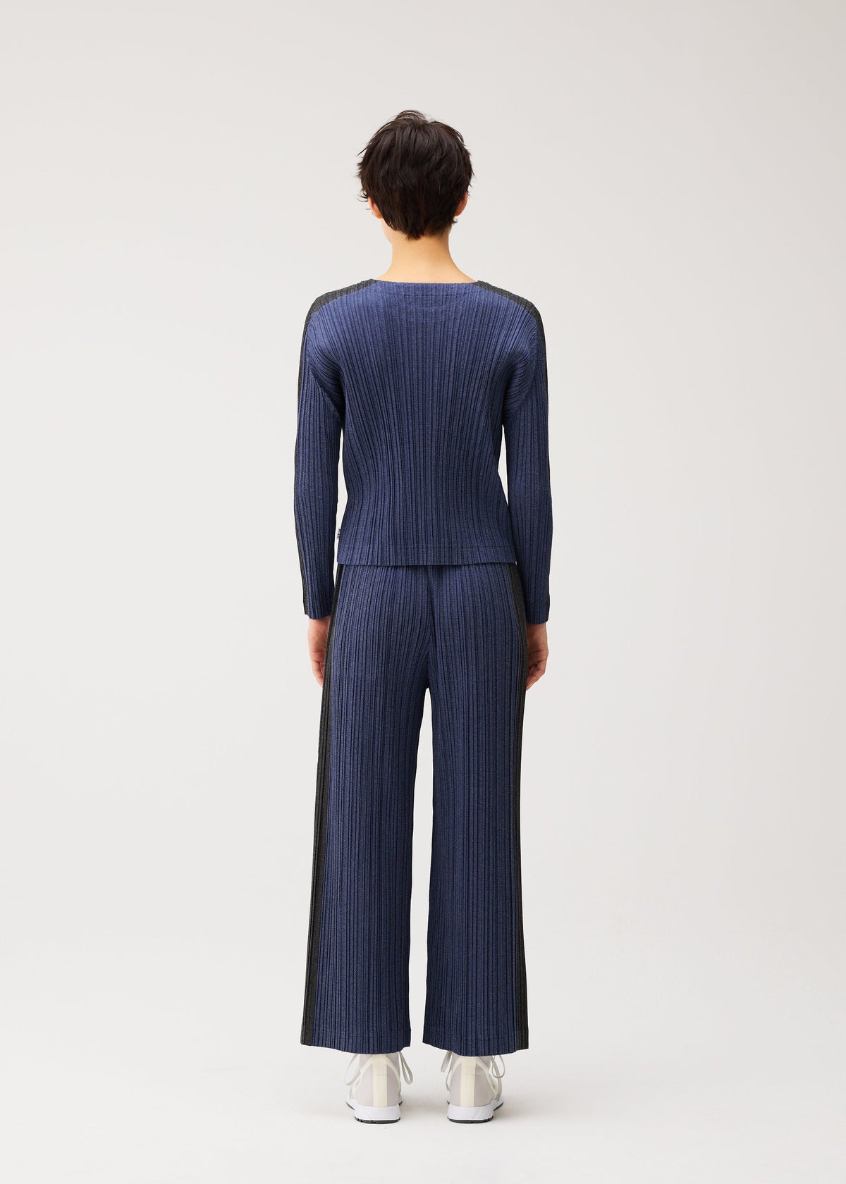 A person stands with their back to the camera, wearing PLEATS PLEASE ISSEY MIYAKE COTTON DENIM—a matching ribbed navy top and straight pants—paired with white shoes against a plain light background. view 7 of 8
