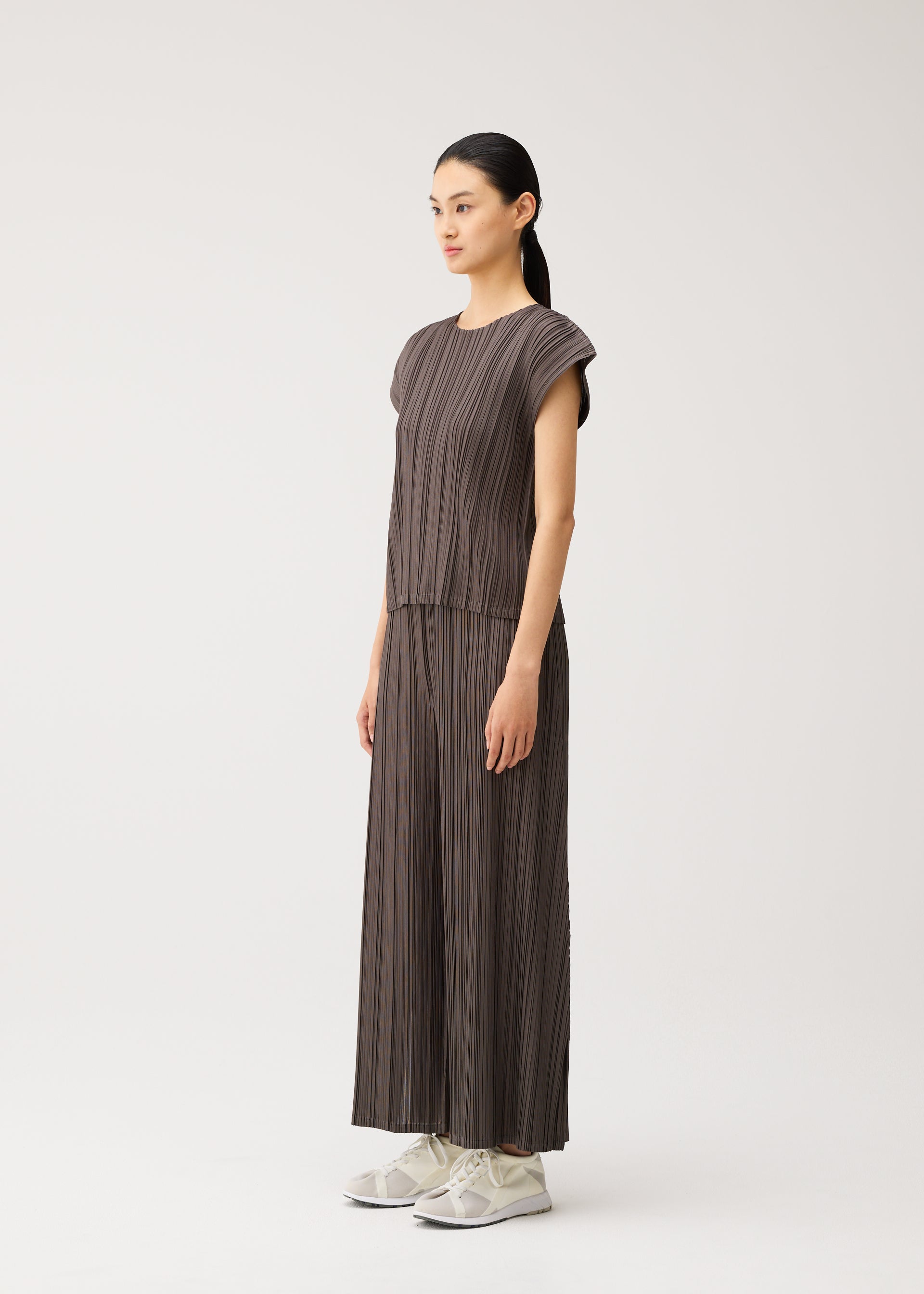 Zoomed view A woman stands in profile against a plain white background, wearing the MELLOW PLEATS set from PLEATS PLEASE ISSEY MIYAKE—a loose, pleated gray top and matching flared pants crafted from mid-weight, soft-textured fabric—paired with white sneakers.