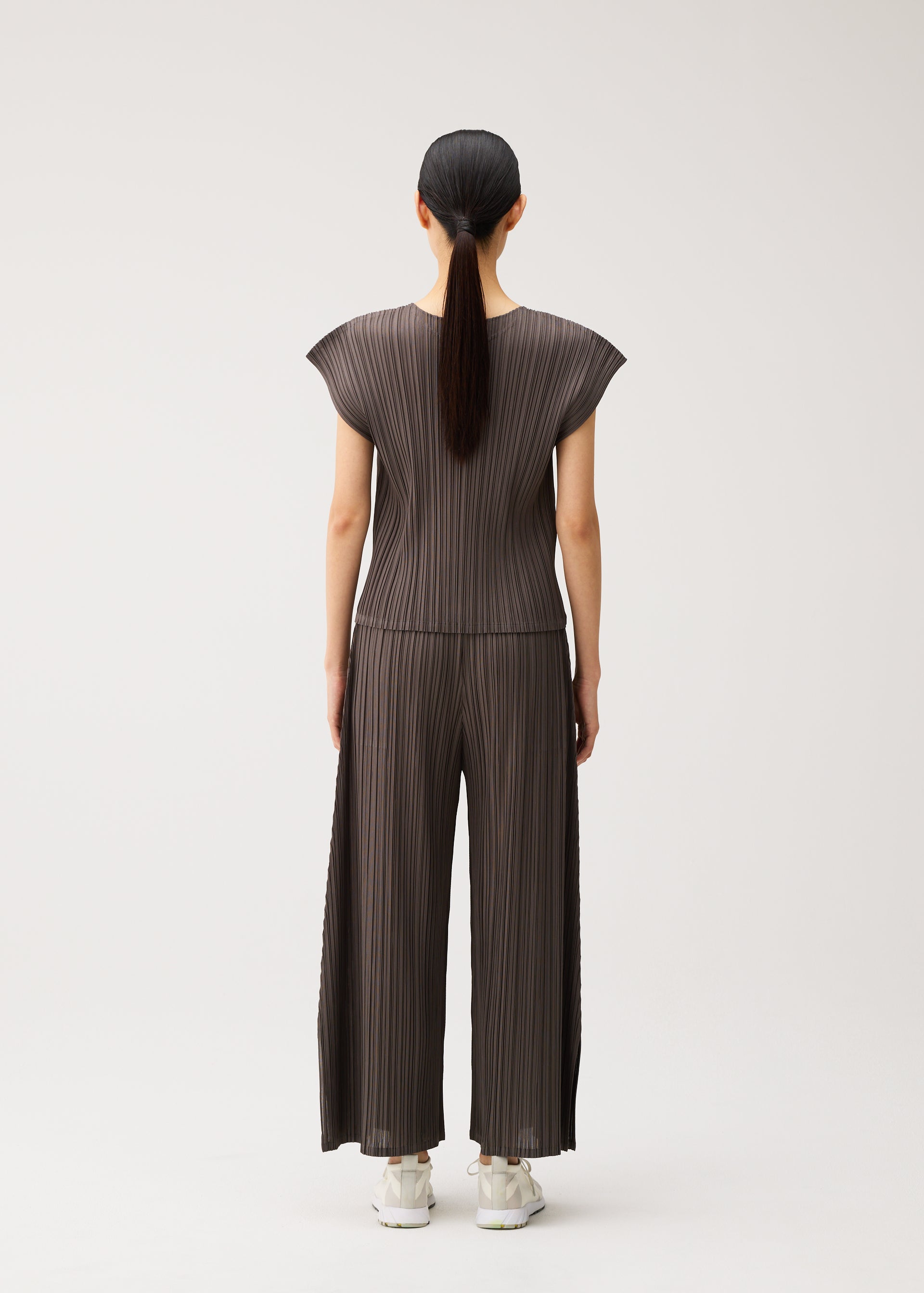 Zoomed view A woman stands facing away, wearing the MELLOW PLEATS brown pleated cap-sleeve top and matching mid-weight flared pants from PLEATS PLEASE ISSEY MIYAKE. Her straight dark hair is tied in a low ponytail, and she pairs the outfit with white sneakers against a plain light background.