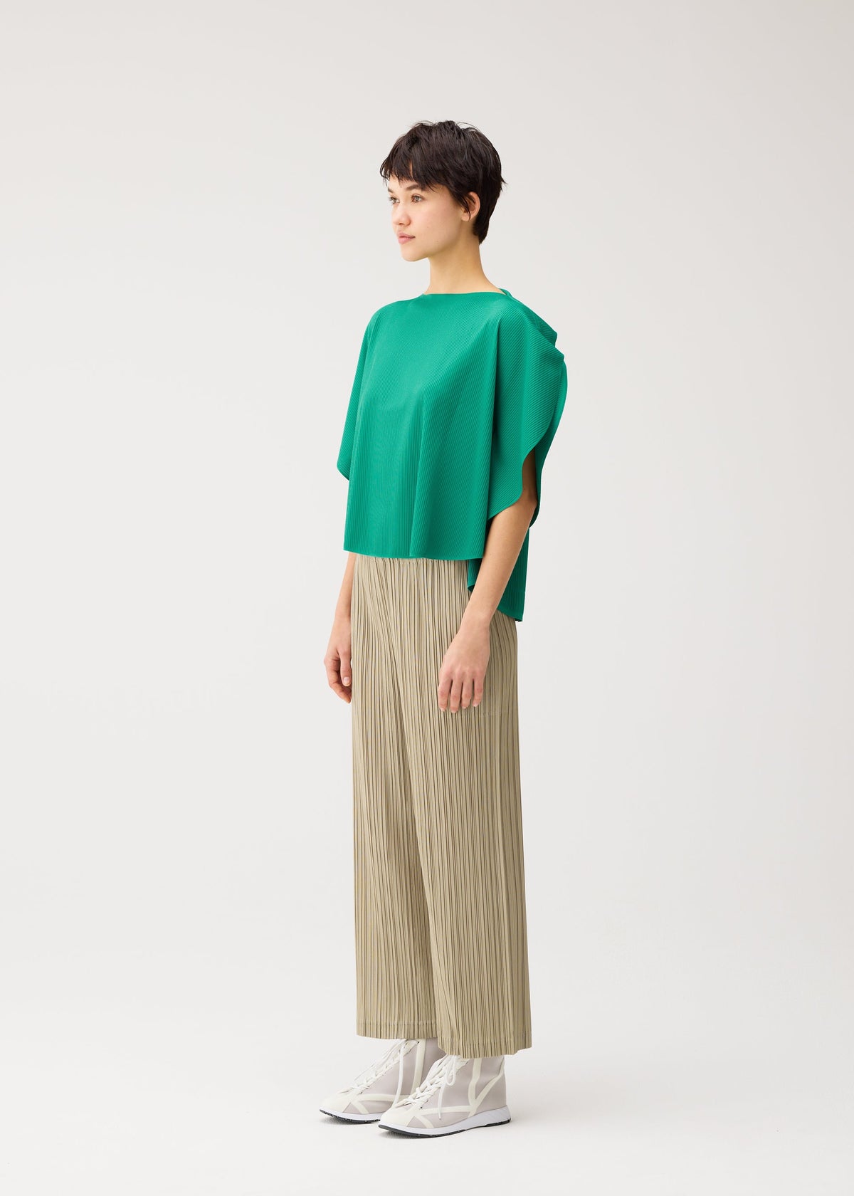 A person stands in profile against a plain background, wearing PLEATS PLEASE ISSEY MIYAKE THICKER BOTTOMS 1—beige mid-weight pleated straight pants—paired with a loose green top and white lace-up shoes. view 8 of 10