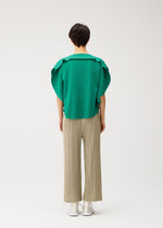 A person faces away from the camera, wearing a green ruffled short-sleeve top and PLEATS PLEASE ISSEY MIYAKE’s THICKER BOTTOMS 1: beige, pleated wide-leg pants in thick fabric, with white shoes, set against a plain white background.