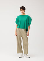 A person with short dark hair and a relaxed expression stands against a plain background, wearing PLEATS PLEASE ISSEY MIYAKE's THICKER BOTTOMS 1 in beige, paired with a loose green top and white sneakers.