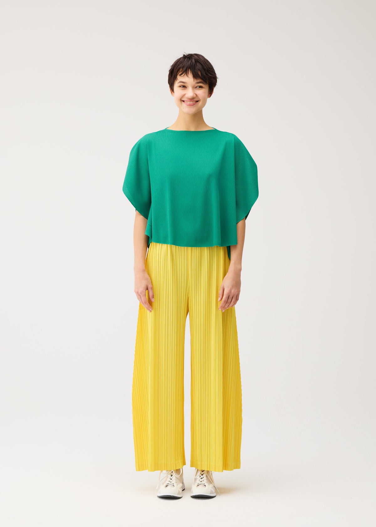 A person stands smiling against a plain white background, wearing a loose green top with pockets, white sneakers, and THICKER BOTTOMS 1 wide yellow pleated pants by PLEATS PLEASE ISSEY MIYAKE. view 7 of 10