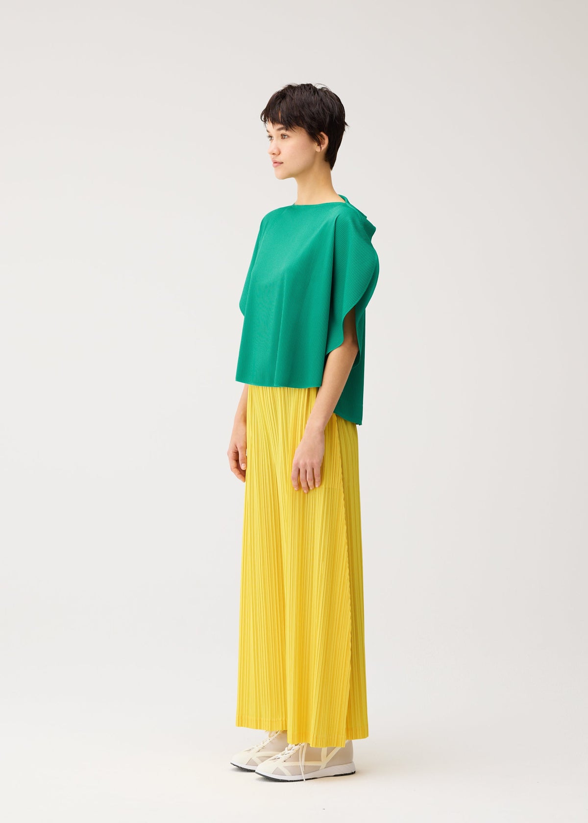 A person stands in profile against a plain white background, wearing PLEATS PLEASE ISSEY MIYAKE THICKER BOTTOMS 1: bright yellow pleated wide-leg pants with pockets, paired with a loose green top and white shoes. view 8 of 10