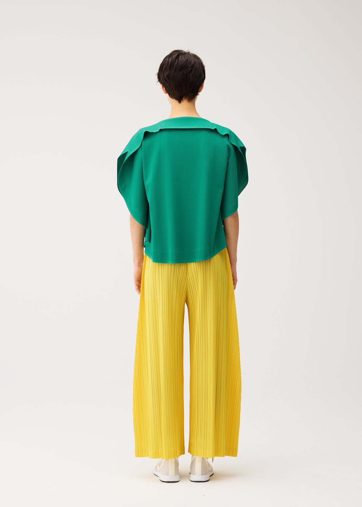 A person stands with their back to the camera, wearing a green short-sleeved top and PLEATS PLEASE ISSEY MIYAKE THICKER BOTTOMS 1 wide yellow pleated pants with pockets, along with white shoes, against a plain light background. view 9 of 10