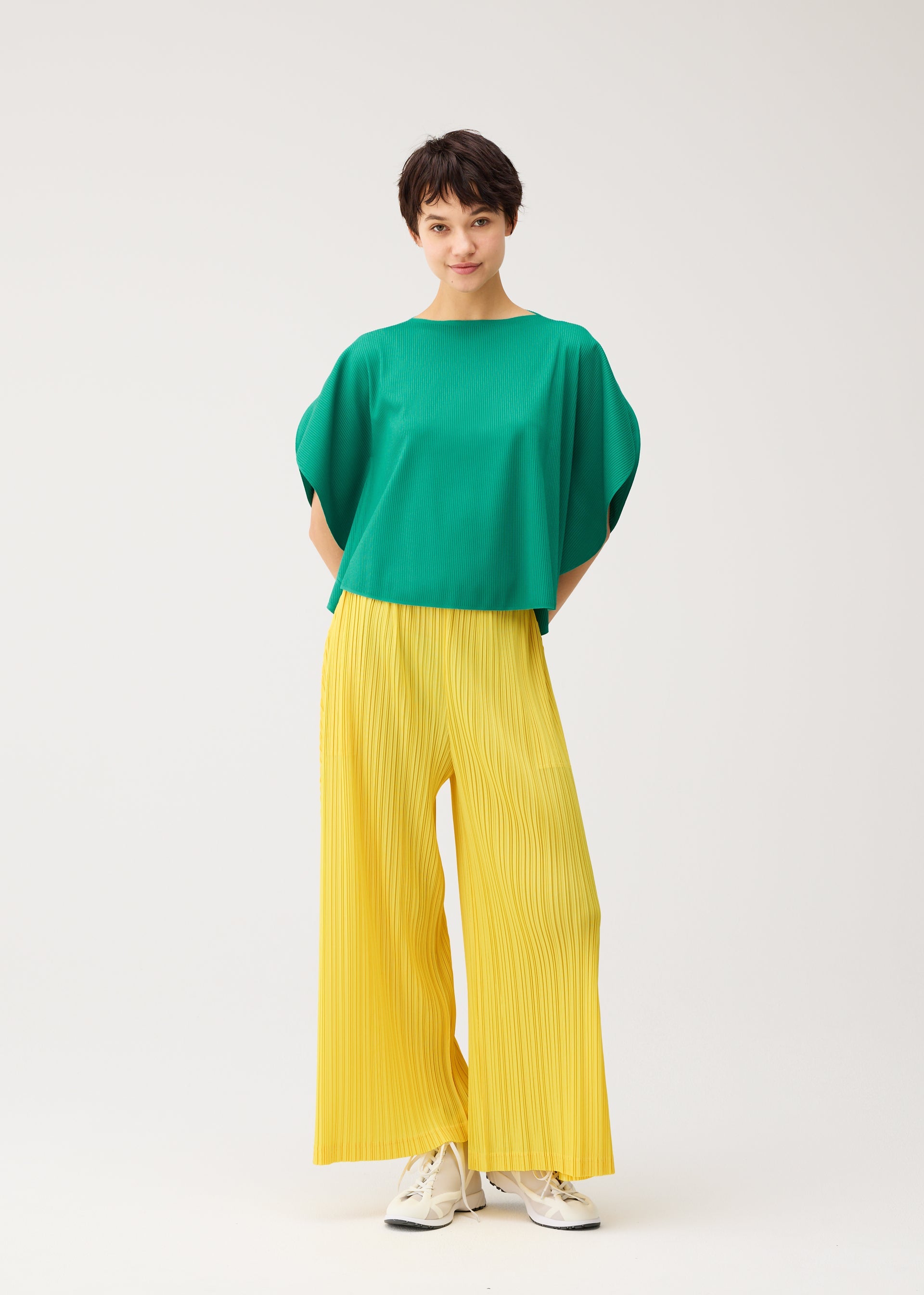 Zoomed view A person in a plain white setting wears PLEATS PLEASE ISSEY MIYAKE’s THICKER BOTTOMS 1—wide yellow pleated pants with pockets and thick fabric—paired with a loose green top, white sneakers, and short dark hair, smiling softly.