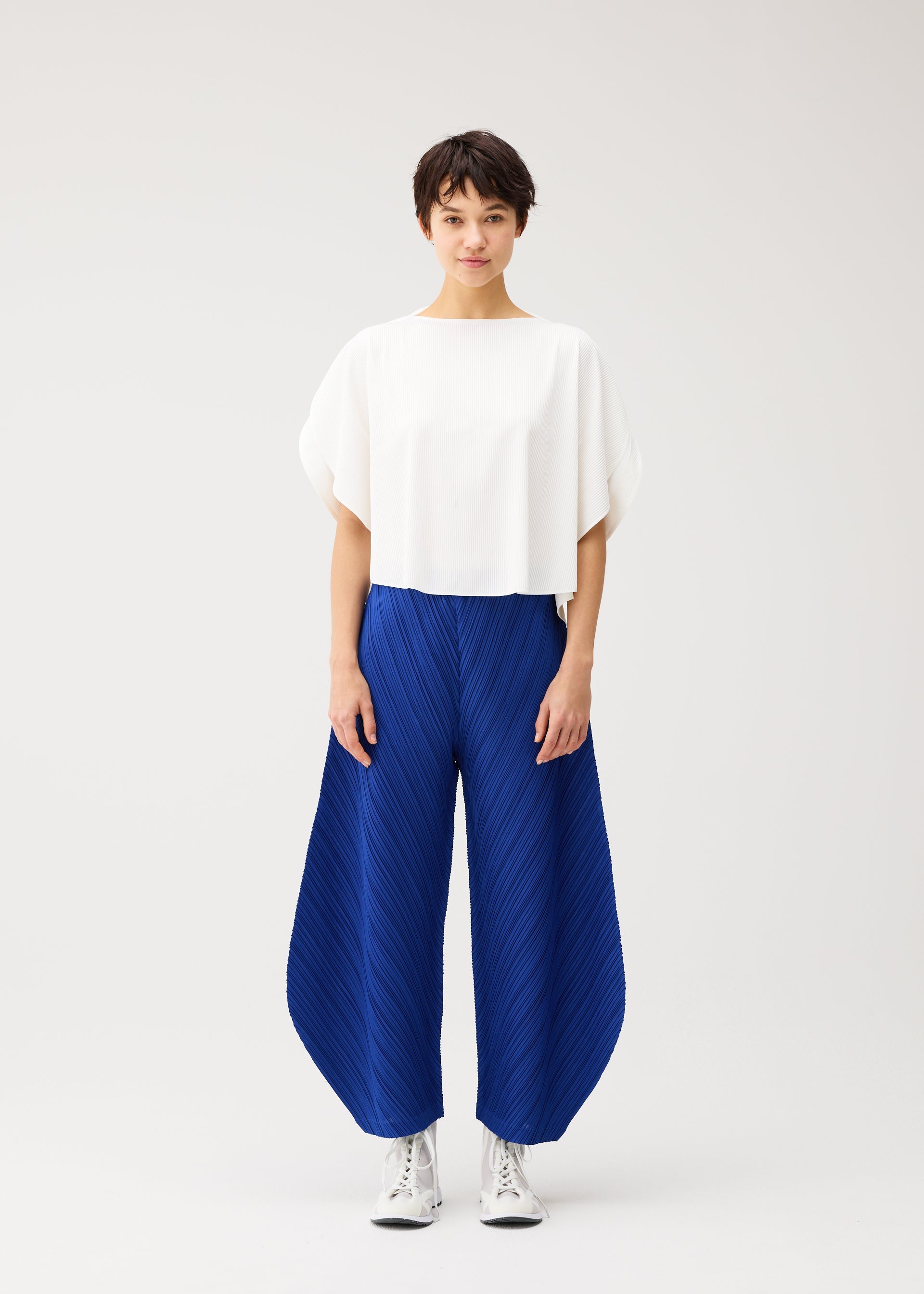 Zoomed view A person with short dark hair wears a loose white top and PLEATS PLEASE ISSEY MIYAKE's THICKER BOTTOMS 1—wide, irregular-cut blue pants with pockets—paired with white lace-up shoes, standing against a plain light background.