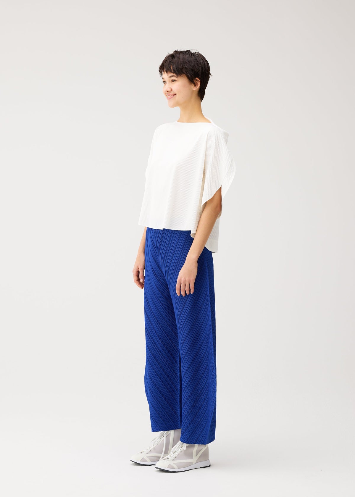 A person stands and smiles, wearing a loose white top, PLEATS PLEASE ISSEY MIYAKE THICKER BOTTOMS 1 bright blue pleated wide-leg pants with pockets, and white shoes, set against a plain light background. view 8 of 10