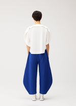 A person with short dark hair, facing away, wears a white top and PLEATS PLEASE ISSEY MIYAKE’s THICKER BOTTOMS 1: wide, irregular-cut blue pants in thick fabric tapering at the ankles, paired with white shoes against a plain light background.