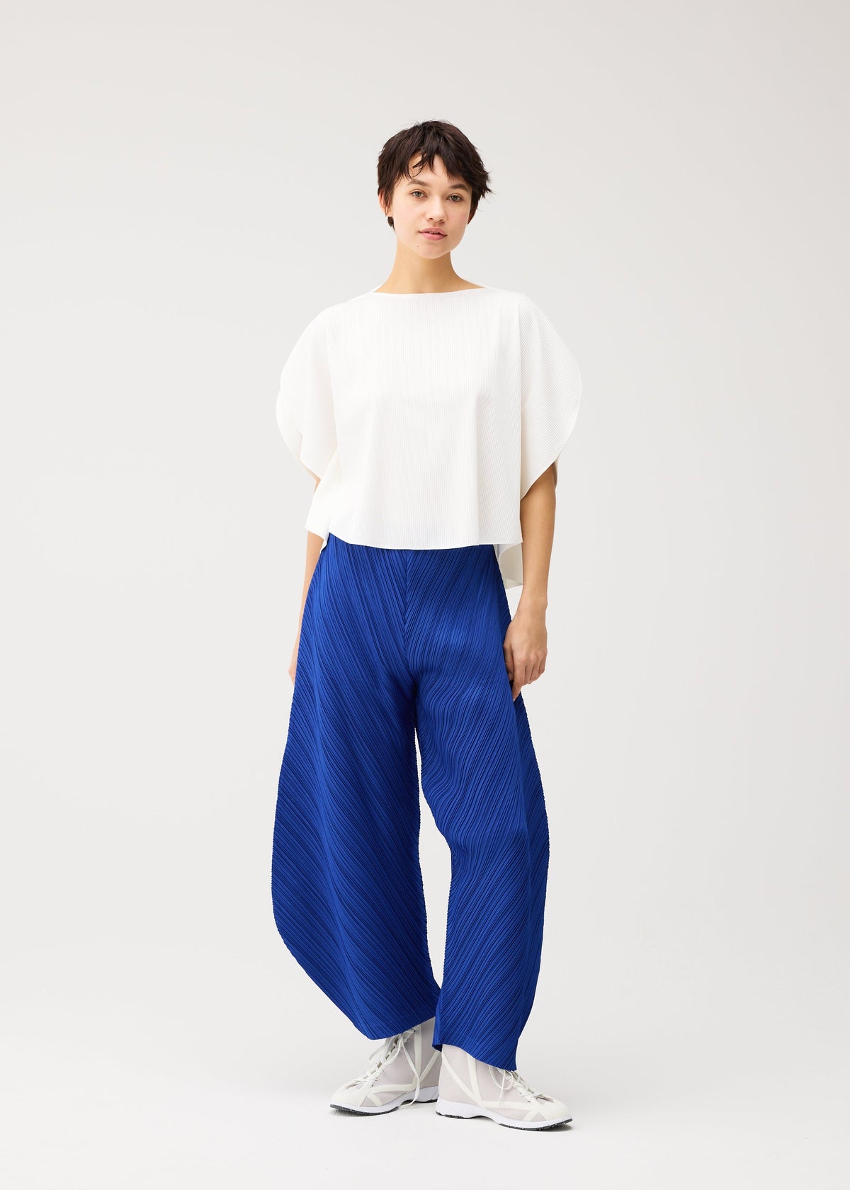 A person stands against a white background wearing THICKER BOTTOMS 1, bright blue pleated wide-leg pants with pockets from PLEATS PLEASE ISSEY MIYAKE, paired with a loose white top and white shoes. view 10 of 10