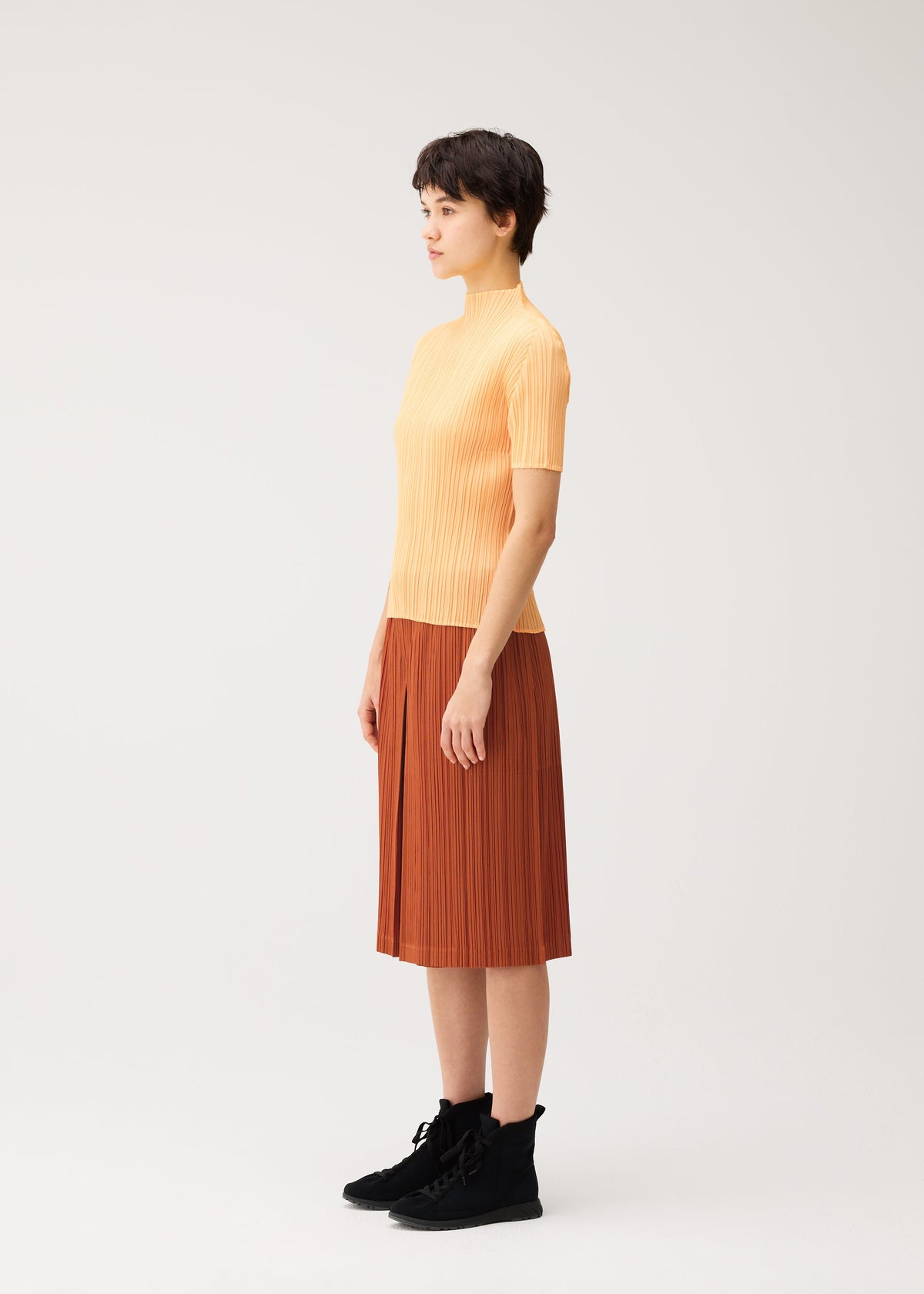 A person stands in profile against a plain background, wearing a short-sleeve yellow top, black boots, and the THICKER BOTTOMS 2 rust-colored knee-length skirt with convenient side pockets by PLEATS PLEASE ISSEY MIYAKE. view 6 of 8