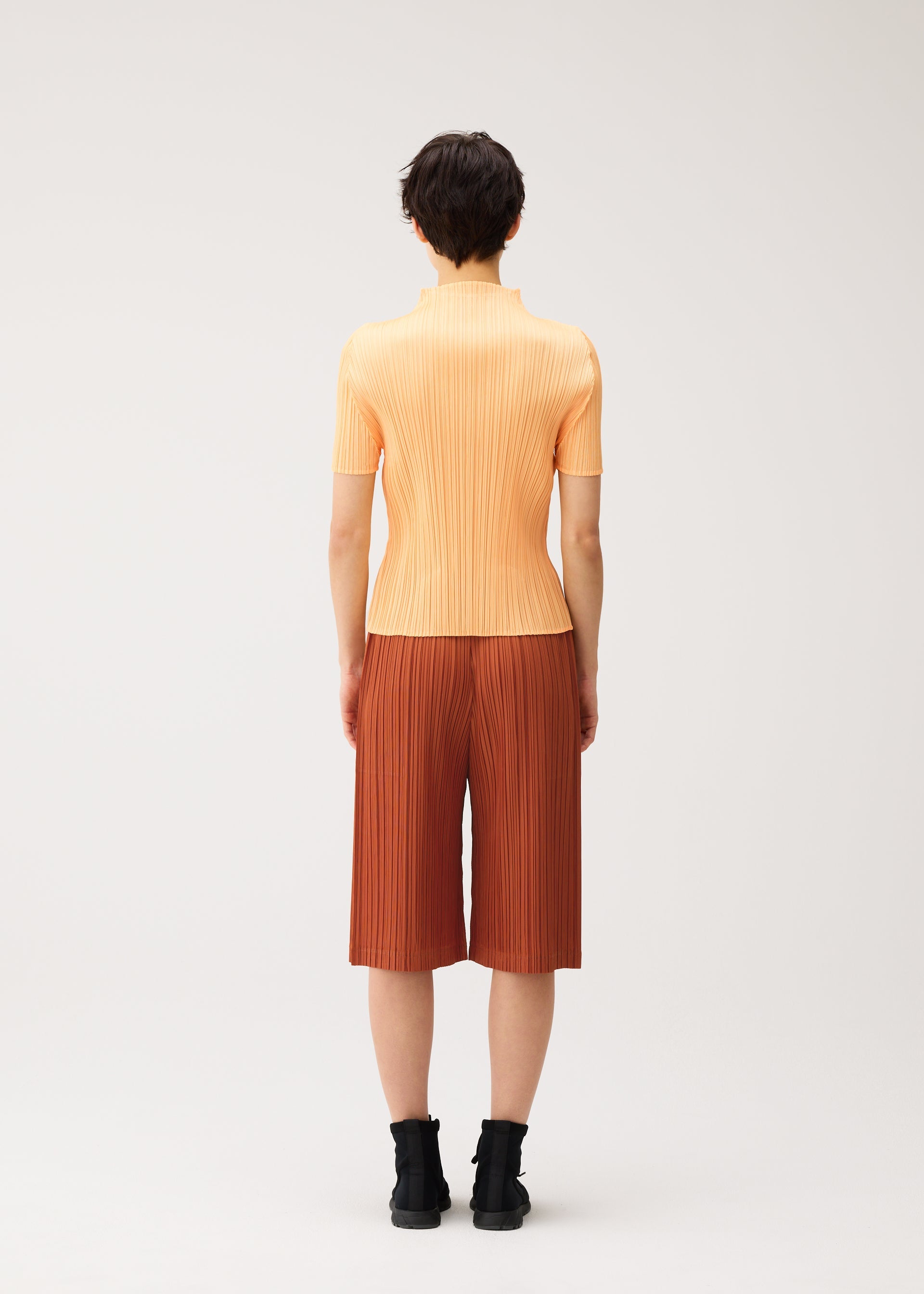 Zoomed view A person stands facing away, dressed in a short-sleeved, light orange, ribbed top and the THICKER BOTTOMS 2 pleated knee-length shorts from PLEATS PLEASE ISSEY MIYAKE. The rust-colored shorts are crafted from thick fabric and feature practical side pockets. They have short dark hair and wear black ankle boots, all set against a plain light background.