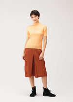 A person wearing the THICKER BOTTOMS 2 rust-colored pleated culottes with side pockets from PLEATS PLEASE ISSEY MIYAKE stands against a plain light background, paired with a short-sleeve, high-neck yellow top and black ankle boots. They smile slightly with a relaxed posture.