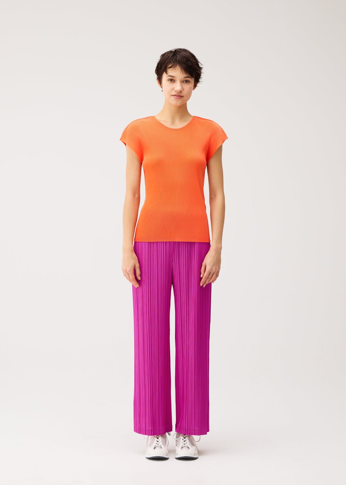 A person stands against a plain background, looking forward with a neutral expression, wearing an orange short-sleeve top and the THICKER BOTTOMS 2 by PLEATS PLEASE ISSEY MIYAKE—bright magenta pleated straight pants crafted from thick fabric with pockets—paired with white sneakers. view 6 of 8