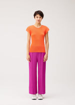 A person stands against a plain background, looking forward with a neutral expression, wearing an orange short-sleeve top and the THICKER BOTTOMS 2 by PLEATS PLEASE ISSEY MIYAKE—bright magenta pleated straight pants crafted from thick fabric with pockets—paired with white sneakers.