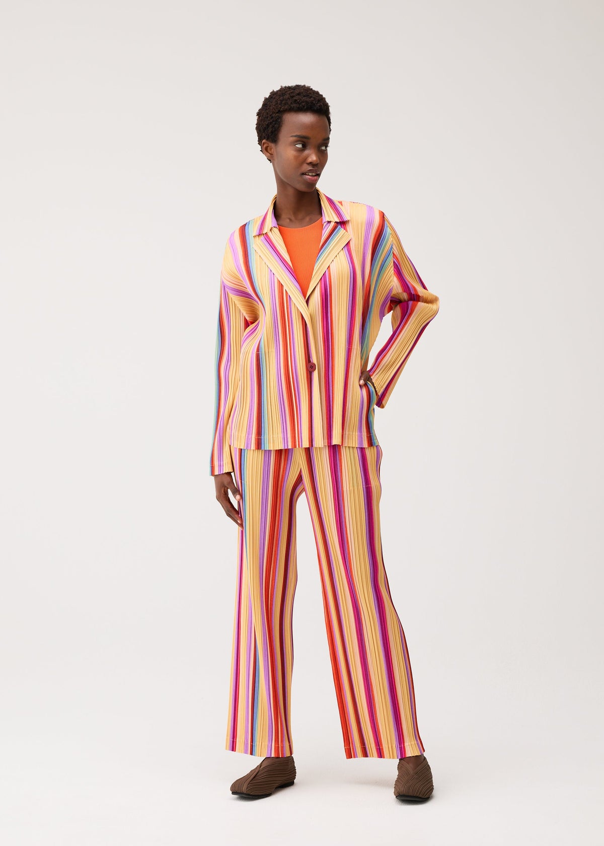 A person models the TAPE TAPE two-piece set by PLEATS PLEASE ISSEY MIYAKE, featuring vertical multicolored stripes and straight-leg pants crafted from thick pleated fabric. They are styled with brown shoes, standing against a plain light background, gazing to the side with one hand on their hip. view 7 of 7