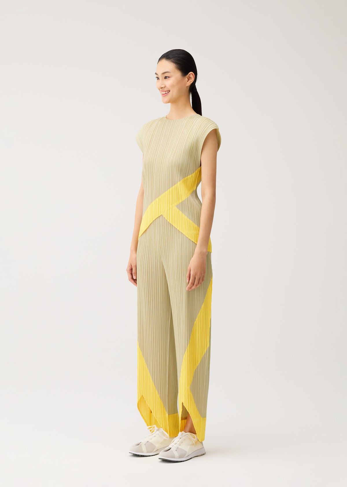 A woman wears the TWIRL outfit by PLEATS PLEASE ISSEY MIYAKE: a beige pleated ensemble with bold yellow diagonal stripes, featuring wrap-style pants inspired by norimaki sushi and convenient side pockets. She completes the look with white sneakers against a simple light background. view 7 of 10