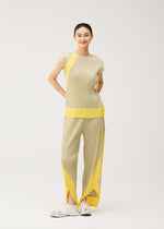 A person stands smiling, dressed in the TWIRL set from PLEATS PLEASE ISSEY MIYAKE—a light beige pleated top and matching wrap-style pants featuring bold yellow geometric accents and handy side pockets—paired with white sneakers against a plain light background.
