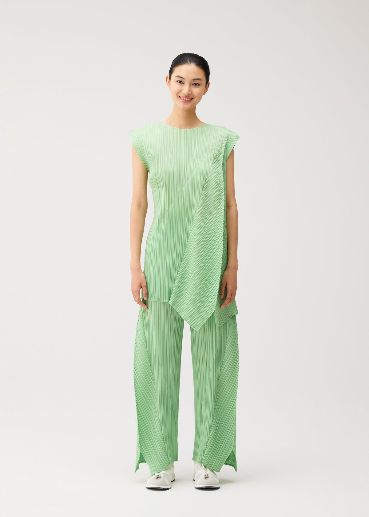A woman stands smiling in the FOLD sleeveless, light green pleated top with matching wide-leg, flared pants featuring pockets by PLEATS PLEASE ISSEY MIYAKE, paired with white sneakers against a plain white background. view 6 of 9