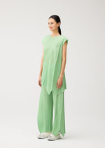 A person stands against a plain white background wearing the FOLD set by PLEATS PLEASE ISSEY MIYAKE—a light green pleated top with matching flared pants, featuring a unique design, subtle pockets, and paired with white shoes.