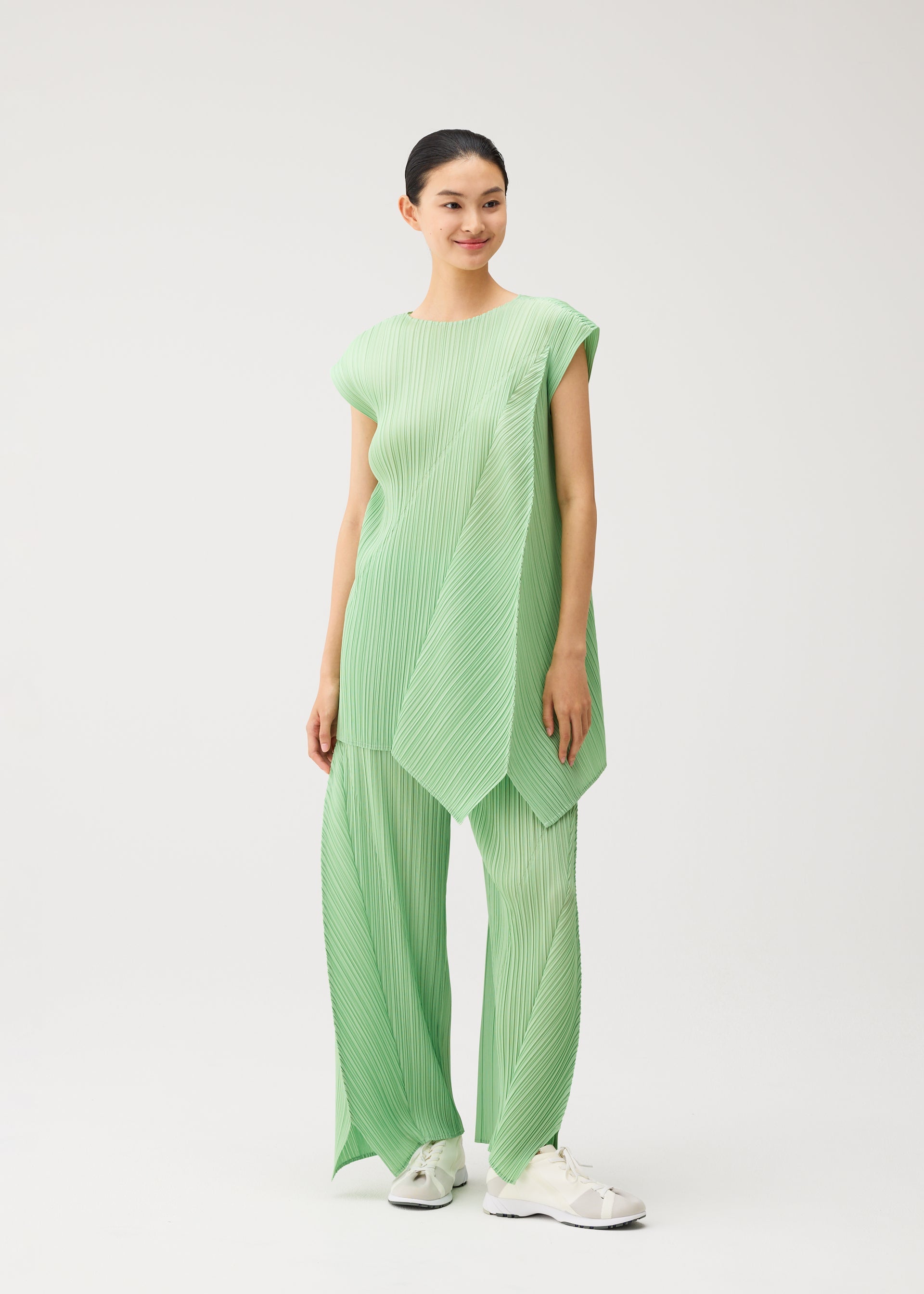 Zoomed view A person wearing the FOLD set by PLEATS PLEASE ISSEY MIYAKE—featuring a light green pleated tunic with unique design details and matching wide-leg, flared pants—stands smiling and looking slightly to the side. They have short, dark hair tied back and are wearing white sneakers, with a plain white background behind them.