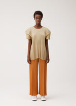 A person faces forward against a light background wearing the BOUQUET set by PLEATS PLEASE ISSEY MIYAKE: a loose, pleated beige top with pockets and wide, pleated orange pants, paired with white sneakers. They have short hair and a neutral expression.