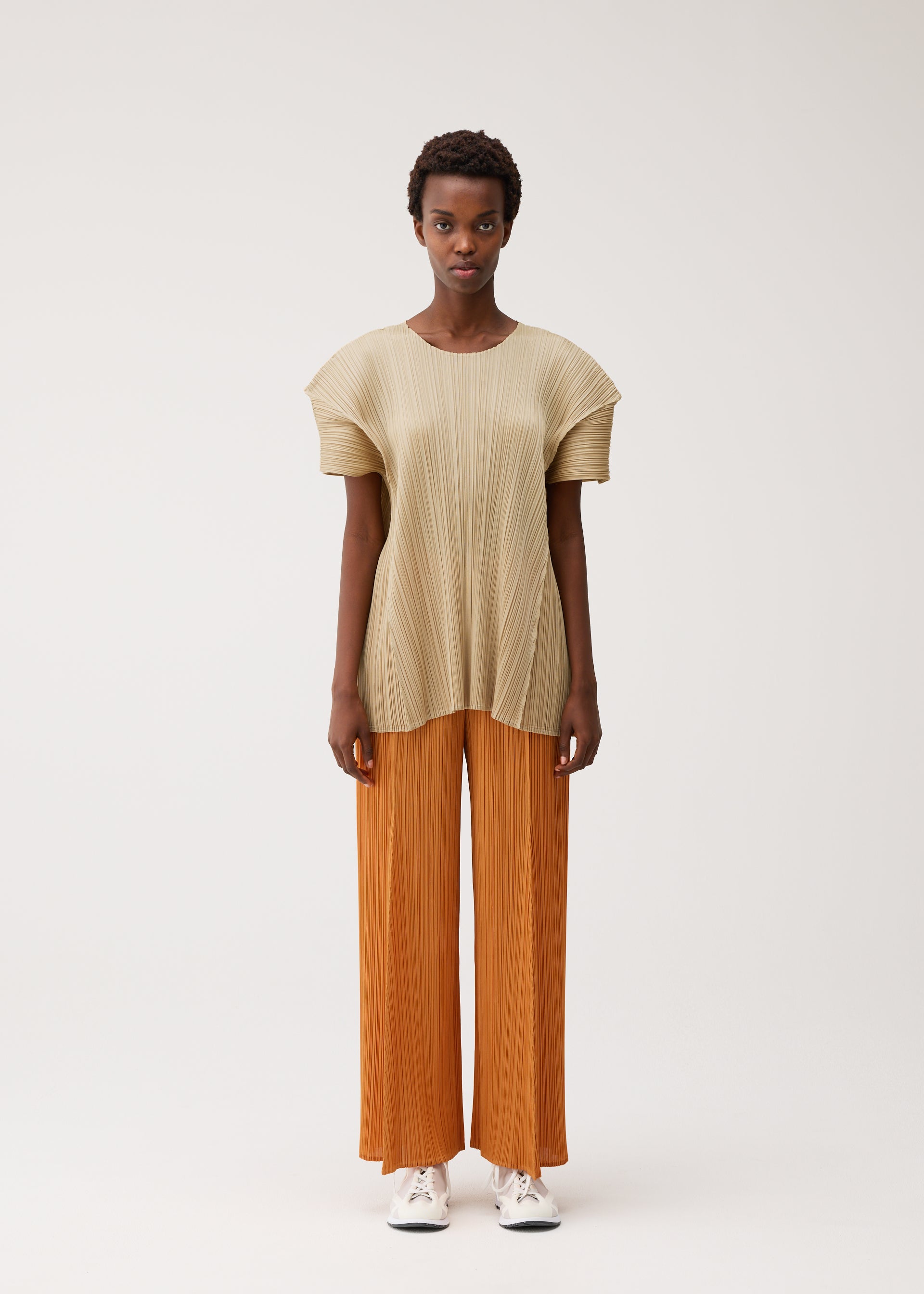 Zoomed view A person faces forward against a light background wearing the BOUQUET set by PLEATS PLEASE ISSEY MIYAKE: a loose, pleated beige top with pockets and wide, pleated orange pants, paired with white sneakers. They have short hair and a neutral expression.