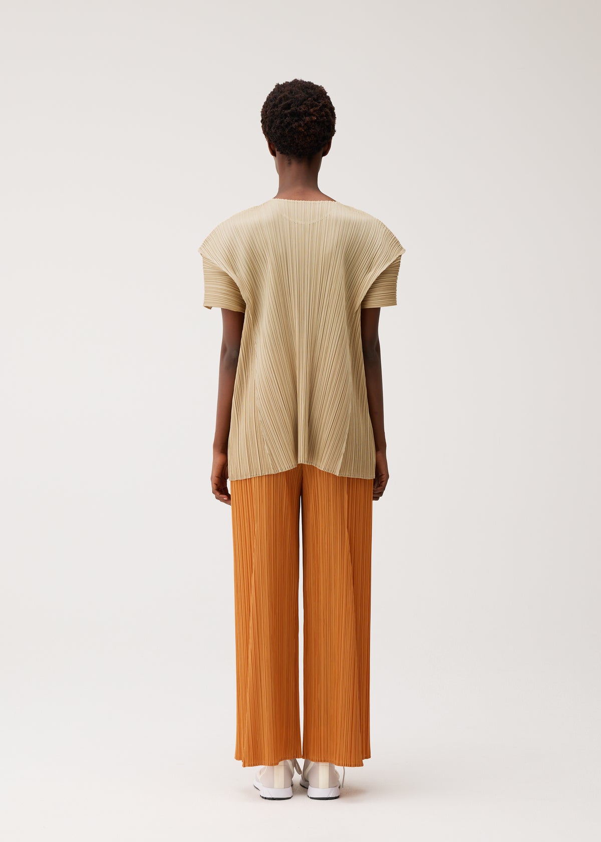 A person stands facing away, wearing the BOUQUET pleated short-sleeve top and wide-leg pants by PLEATS PLEASE ISSEY MIYAKE. The outfit is beige and orange with pockets; they wear white shoes. The background is plain and light-colored. view 7 of 8