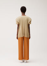 A person stands facing away, wearing the BOUQUET pleated short-sleeve top and wide-leg pants by PLEATS PLEASE ISSEY MIYAKE. The outfit is beige and orange with pockets; they wear white shoes. The background is plain and light-colored.