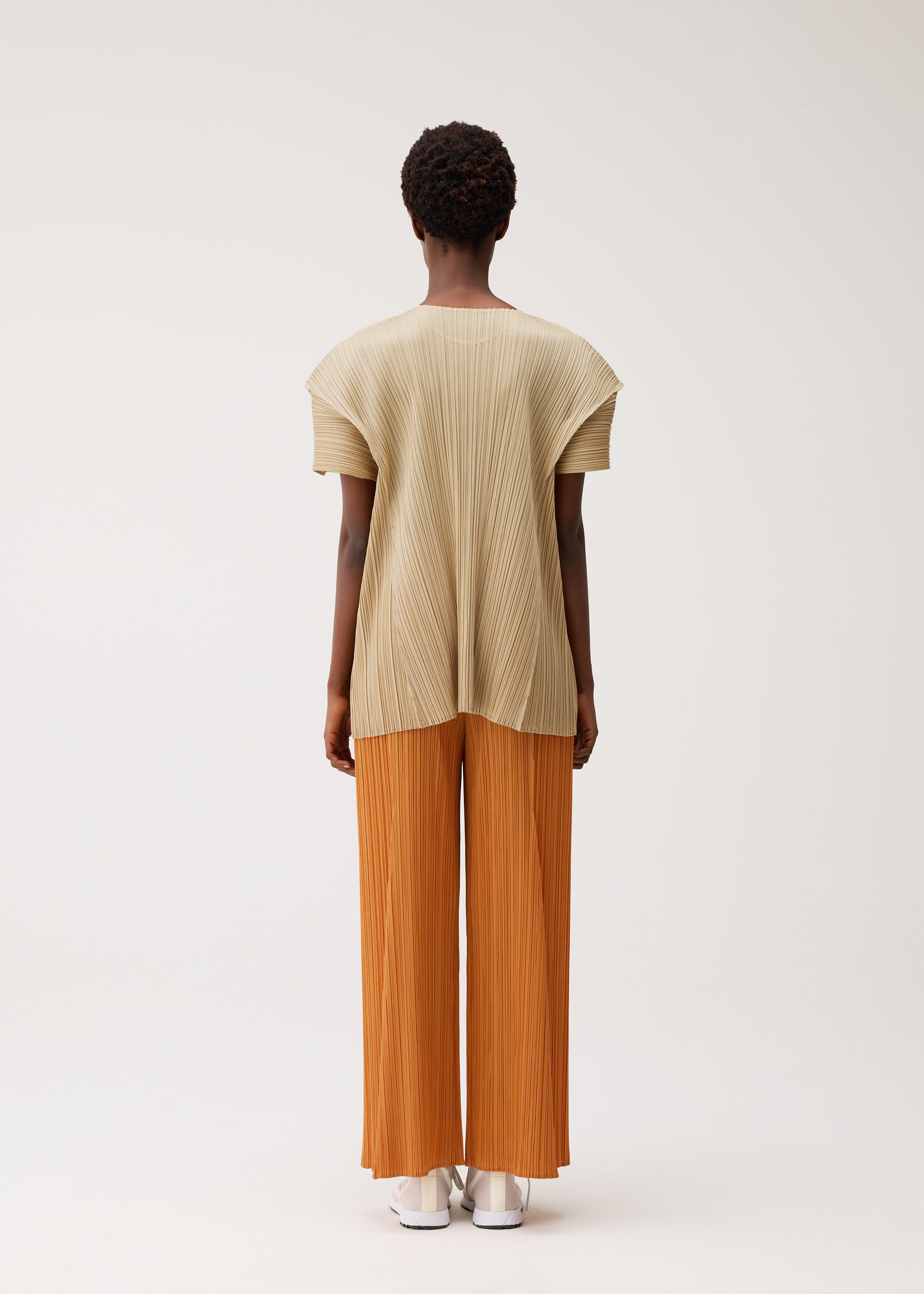 Zoomed view A person stands facing away, wearing the BOUQUET pleated short-sleeve top and wide-leg pants by PLEATS PLEASE ISSEY MIYAKE. The outfit is beige and orange with pockets; they wear white shoes. The background is plain and light-colored.