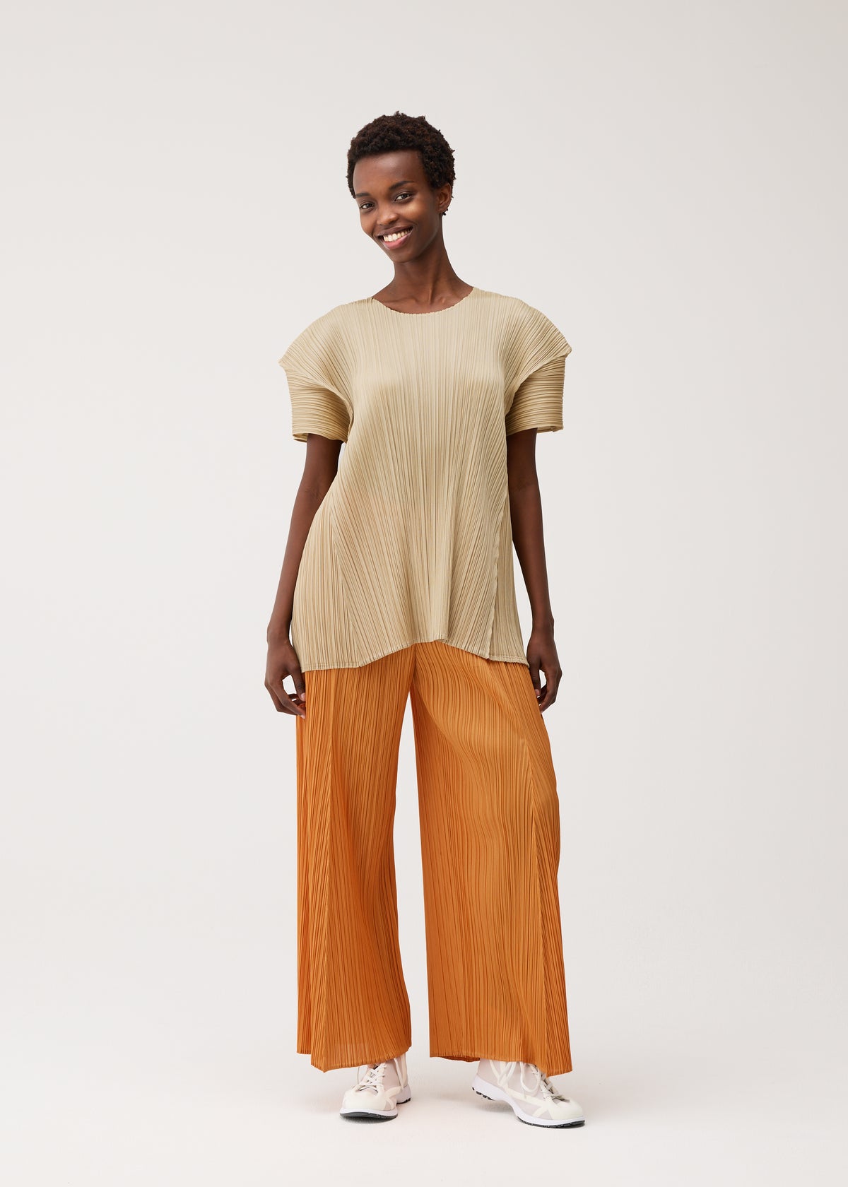 A person smiles holding BOUQUET from PLEATS PLEASE ISSEY MIYAKE, dressed in a beige pleated top, wide orange pleated pants with pockets, and white sneakers, standing against a plain light background. view 8 of 8