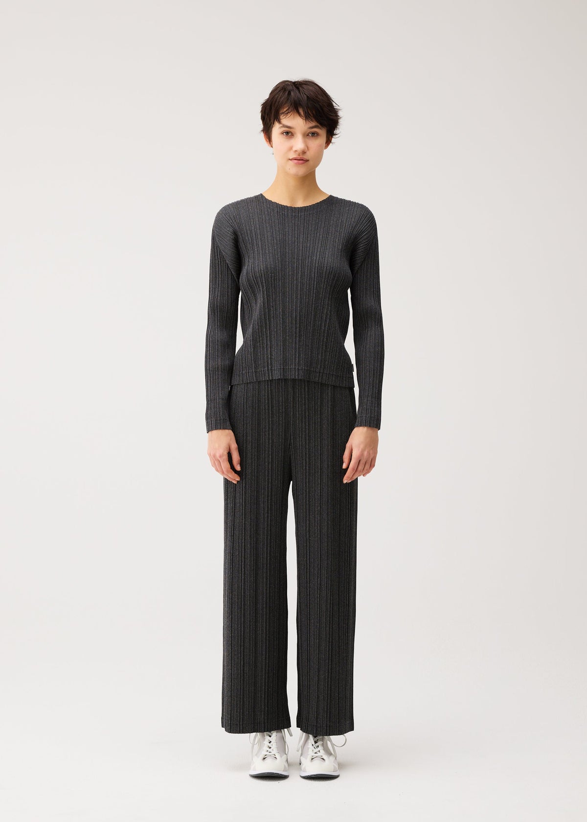 A person stands facing forward in the PLEATS PLEASE ISSEY MIYAKE COTTON DENIM BLACK set—dark, ribbed knit sweater with matching straight pants—and white sneakers, against a light background. view 3 of 6