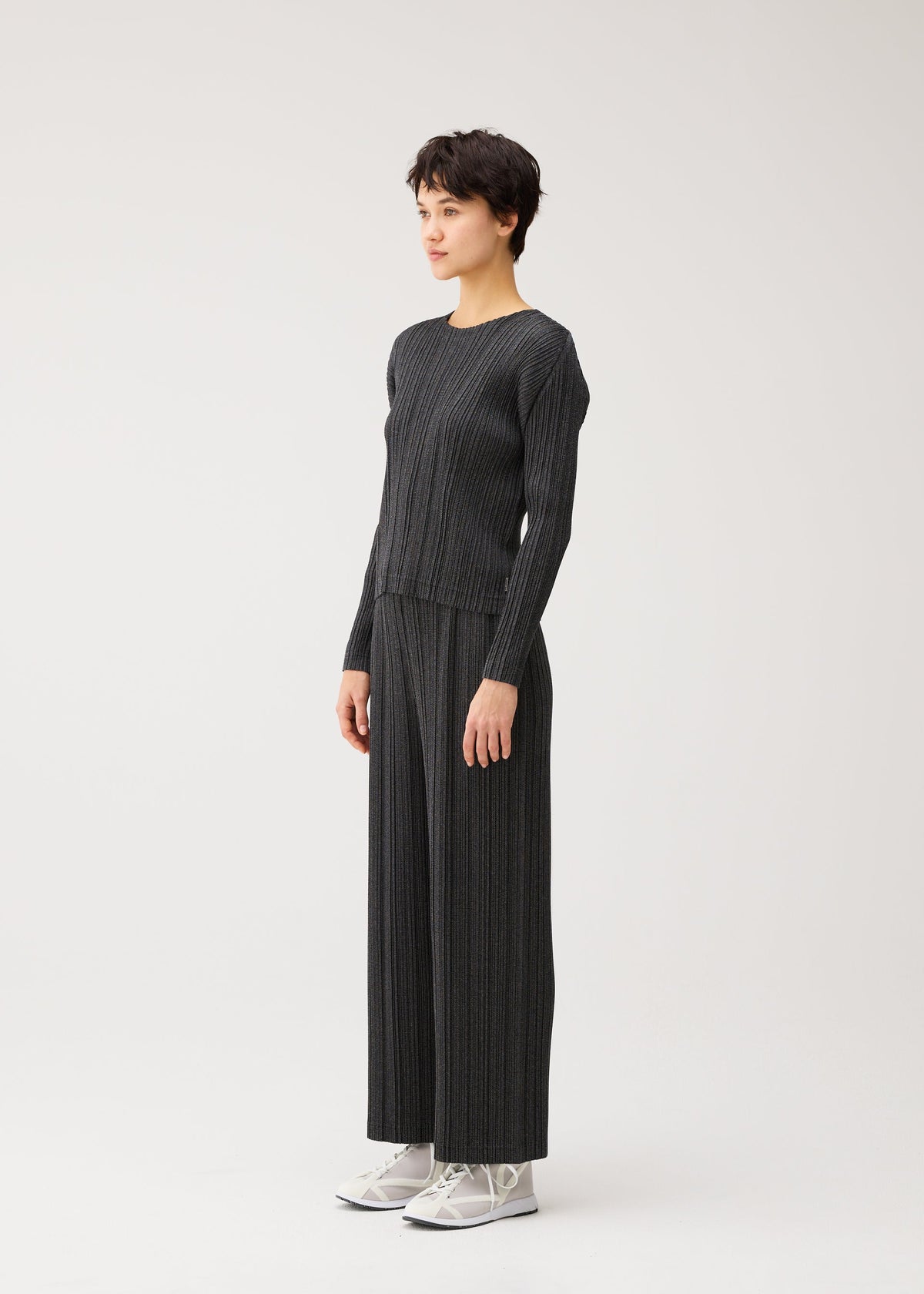 A person stands in a minimalist space wearing PLEATS PLEASE ISSEY MIYAKE’s COTTON DENIM BLACK set with white sneakers, facing slightly to the side and smiling gently. view 4 of 6