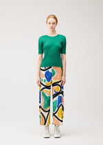 A woman stands against a plain background wearing the ROLL green short-sleeved top and abstract print pants by PLEATS PLEASE ISSEY MIYAKE, paired with white lace-up shoes.