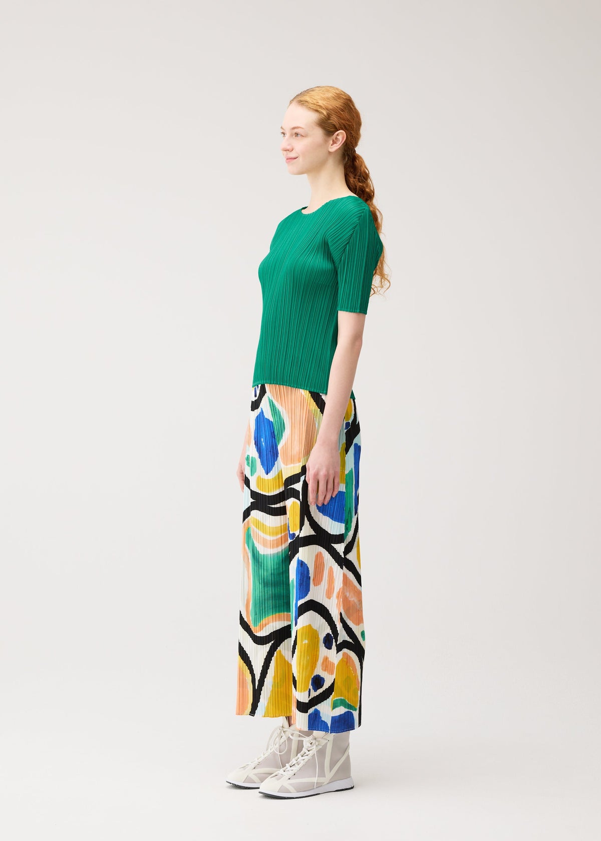 A woman with long red hair in a ponytail stands sideways on a plain background, wearing PLEATS PLEASE ISSEY MIYAKE’s ROLL green short-sleeved top and abstract patterned pants, paired with white high-top sneakers. view 5 of 7