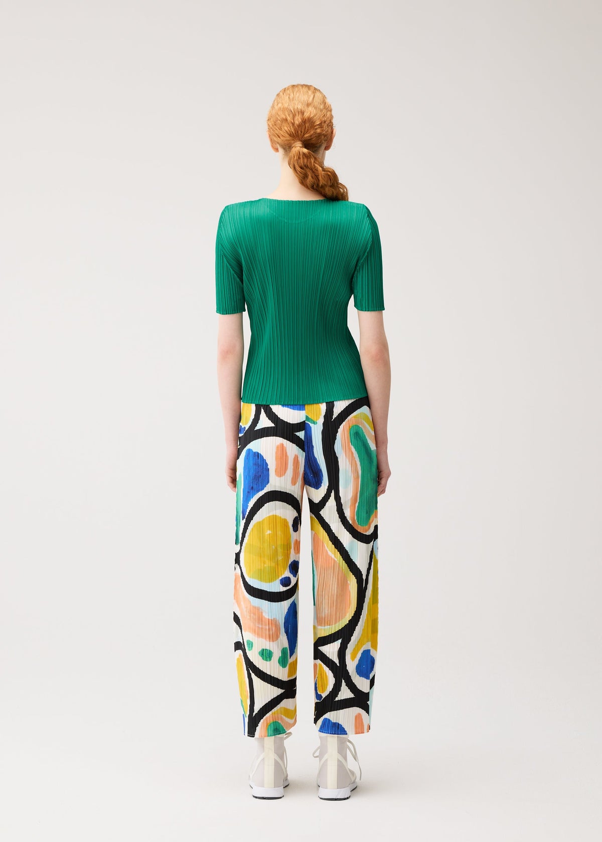 A person with long red hair in a braid wears the PLEATS PLEASE ISSEY MIYAKE ROLL green short-sleeved top and colorful print semi-wide pants, standing facing away against a plain light background. view 6 of 7
